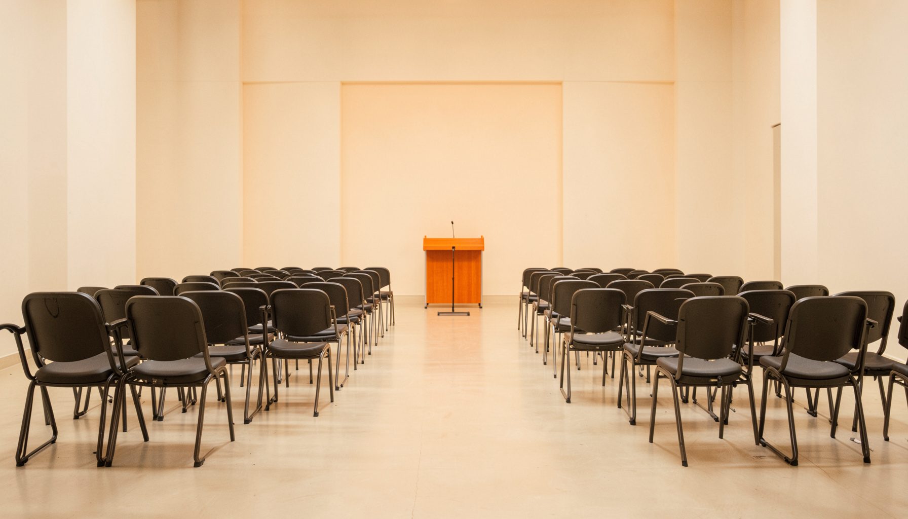 A clean symmetrical photograph of empty rehearsal chairs set in rows with warm off-white walls and a single conductor stand