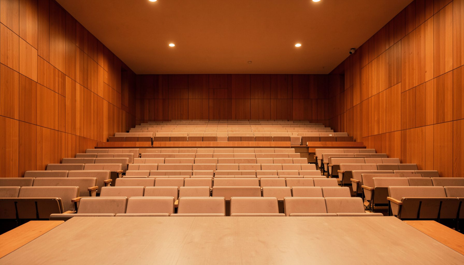 A clean, symmetrical photograph of an empty concert hall interior with warm wooden paneling, soft amber lighting, and tiered seating