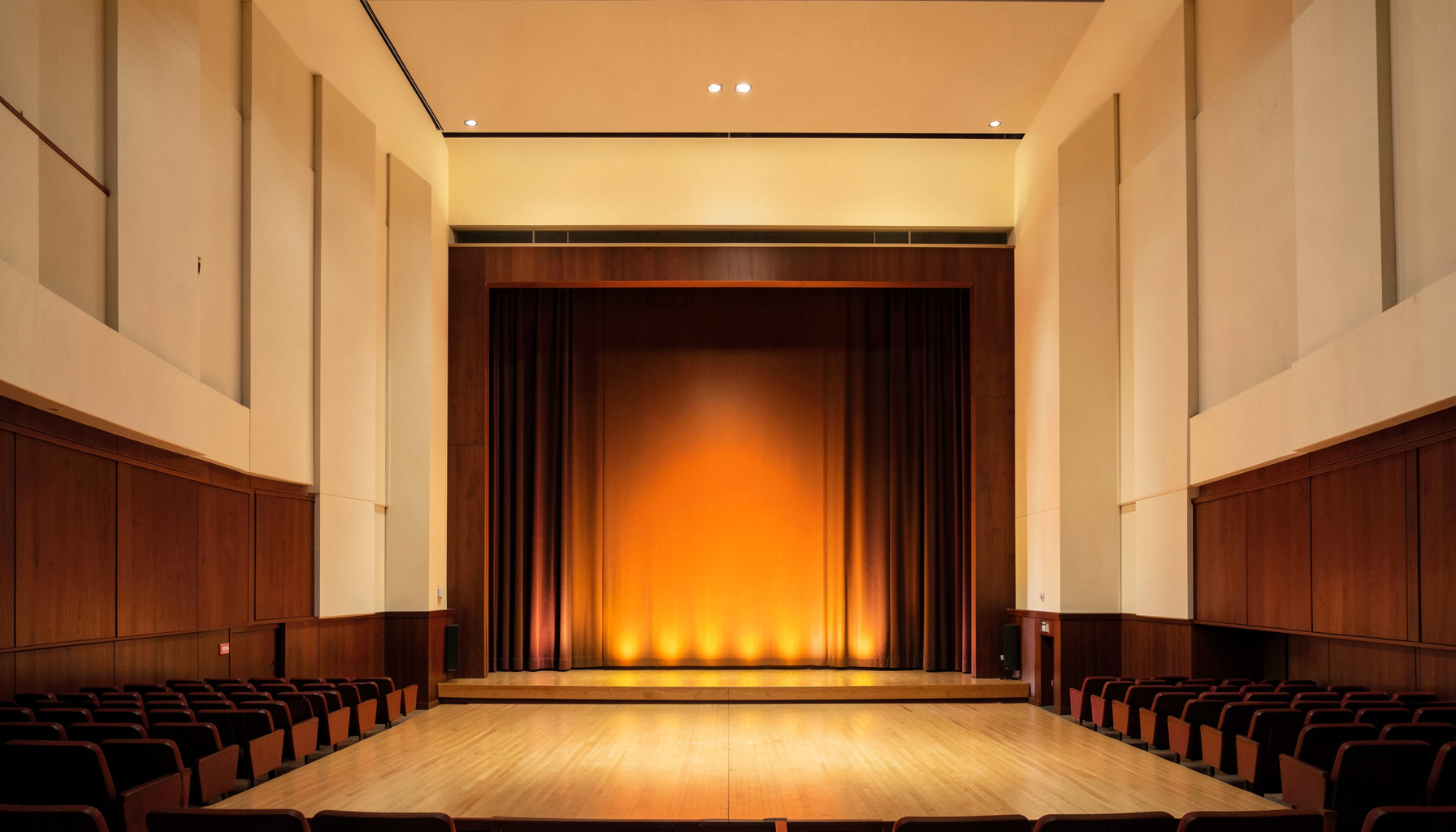 Soft warm neutral concert hall with pale cream walls, deep mahogany wood, and golden stage light